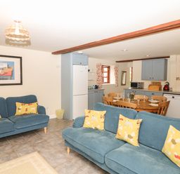 A living room with blue sofas and yellow pillows next to a kitchen with blue cabinets and a wooden dining table at Battens in Colyton