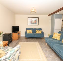 A living room with two blue sofas with yellow cushions a patterned armchair a wooden TV stand with a television and a small black stove at Battens in Colyton