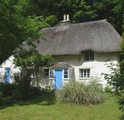 A cottage with a thatched roof blue doors and a garden with plants and trees at Lew Quarry Cottage in Lewdown