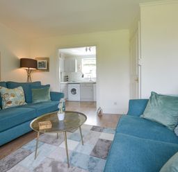 A living room with blue sofas a round brass coffee table and a view into a white kitchen at Linney Cottage in Dittisham