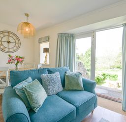 A living room with a blue sofa and cushions a dining table with chairs and flowers a large wall clock and glass doors with curtains at Linney Cottage in Dittisham