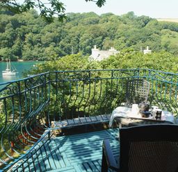 A balcony with two chairs and a table overlooking a river with a boat and trees at Galanda Treetops in Newton Ferrers