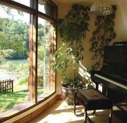 A room with a black piano and a stool next to a large window overlooking a garden and trees at Galanda Treetops in Newton Ferrers