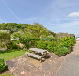 A garden area with a wooden picnic table on a stone patio surrounded by trees and bushes at 4 Coastguards Cottage in Hope Cove