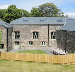 An outdoor area with stone buildings trees and a wooden fence surrounding a patio with a table and chairs at Mill Barn in Woolston near Salcombe