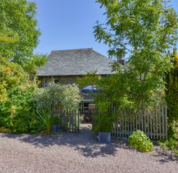 A garden with trees and bushes surrounding a wooden fence and stone house at Parsonage Farm Cottage in Newton Ferrers
