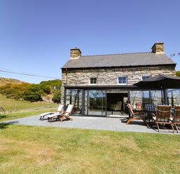 A stone farmhouse with a slate roof and a patio with wooden chairs and a large umbrella at Garth Morthin The Farmhouse in Porthmadog