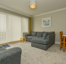 A living room with grey sofas a small wooden table vertical blinds and a dining table with chairs at 3 Tarifa in Bigbury-on-Sea