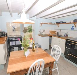 A kitchen with wooden table and two chairs a stove microwave and sink with shelves holding dishes at The Bathing House in Ashprington