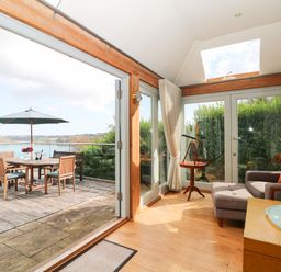 A seating area with a chair ottoman and telescope next to glass doors opening to a wooden deck with a dining table and umbrella at Dove Cottage in Dittisham