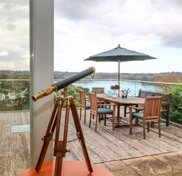 A telescope on a wooden tripod inside looking out to a wooden deck with a dining table and chairs under a large umbrella at Dove Cottage in Dittisham