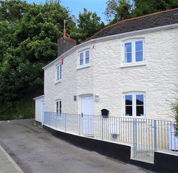 A white stone house with metal railing along the front on a curved road with trees in the background at Junket Cottage in Noss Mayo