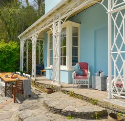 An outdoor patio area with a wooden dining table surrounded by chairs and two cushioned armchairs beside a light blue building with white framed windows at Woodlands in Dartmouth