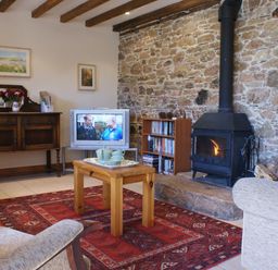 A living room with a wood stove and stone wall a wooden coffee table with a tea set a red patterned rug a television on a stand a wooden chest a bookshelf and exposed ceiling beams at The Clock House in Ivybridge