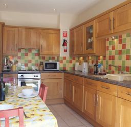 A kitchen with wooden cabinets a tiled multicolored backsplash a table with a yellow floral tablecloth set with plates and glasses at The Clock House in Ivybridge