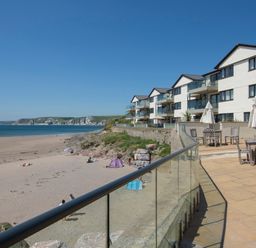 A beachfront patio with tables and umbrellas next to a row of residential buildings overlooking a sandy beach and ocean cliffs at 5 Burgh Island Causeway in Bigbury-on-Sea