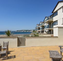 A patio with wooden chairs and tables overlooking the beach and sea with modern buildings on the right at 13 Burgh Island Causeway in Bigbury-on-Sea