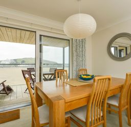 A dining area with a wooden table and chairs next to glass sliding doors leading to a balcony with outdoor furniture at 13 Burgh Island Causeway in Bigbury-on-Sea