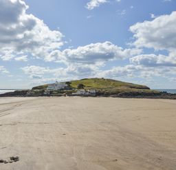 A sandy beach leading to a grass-covered island with buildings under a partly cloudy sky at 24 Burgh Island Causeway in Bigbury-on-Sea