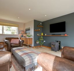 A living room with brown leather armchairs a plaid ottoman a wall-mounted television and a wood stove at Orchard View in Kellaton near Hallsands