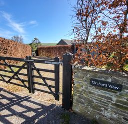 A black wooden gate at the entrance to a gravel driveway with hedges on both sides at Orchard View in Kellaton near Hallsands
