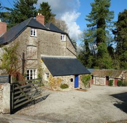 A stone house with a black roof and blue door next to wooden gate and outbuildings surrounded by trees at Maristow Barton in Milton Combe near Lopwell Dam