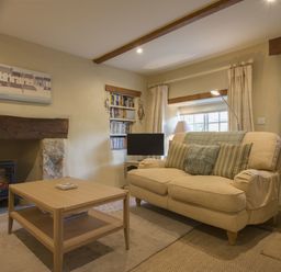 A living room with a beige sofa pillows a wooden coffee table a small fireplace a bookshelf and a window at Cherry Tree Cottage in Noss Mayo