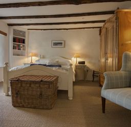 A bedroom with a bed two bedside tables with lamps a wicker chest an armchair and a wooden wardrobe at Cherry Tree Cottage in Noss Mayo