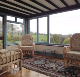 A seating area with three cushioned armchairs and a small table near large windows overlooking a garden at Higher Tumrose in Blisland