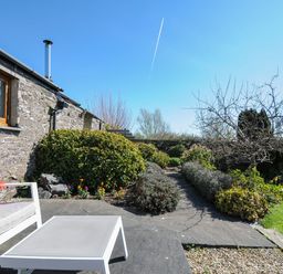 A stone wall with a window and a chimney next to a garden path with bushes and a tree and outdoor seating with a table and chair at Swallow Barn in St Kew