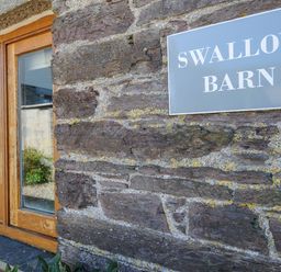 A stone exterior wall with a wooden framed window and a sign that reads Swallow Barn at Swallow Barn in St Kew
