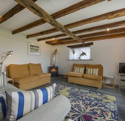 A living room with wooden ceiling beams armchairs a patterned rug a small stove and a television at Salty Cottage in Morwenstow