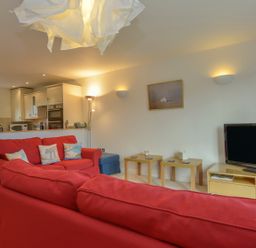 A living room with red sofas two wooden side tables and a television next to a kitchen at The Loft in Bude