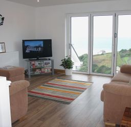 A living room with brown sofas a striped rug a television and large glass doors showing a hill and sea view at Seagulls Perch in Mawgan Porth
