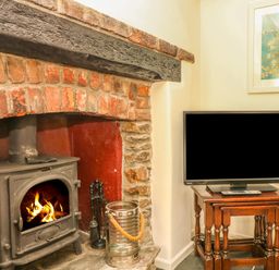 A wood burning stove with fire in a brick fireplace next to a wooden nest of tables with a flat screen tv on top at Cousham Cottage in Kingsand And Cawsand