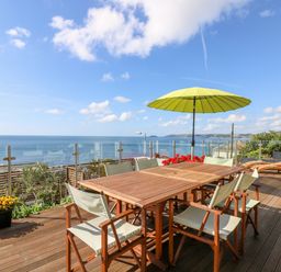 An outdoor wooden deck with dining table and chairs a yellow umbrella and two wooden loungers overlooking the sea at Rock Rose in Downderry