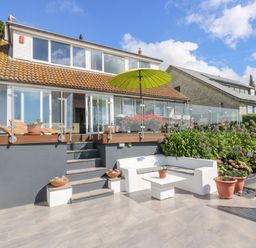 An outdoor patio area with white seating and potted plants in front of a house with glass doors and a green umbrella at Rock Rose in Downderry