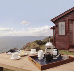 A picnic table with coffee cups, a French press, biscuits, binoculars, and a wooden cabin with an open door by the seaside at Seagulls Nest Whitsand Bay near Millbrook