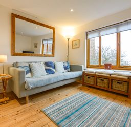 A living room with a sofa cushions a large mirror a wooden side table lamps a window seat with baskets and a striped rug at Chough Cottage in Kingsand And Cawsand