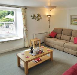 A living room with a beige sofa and red cushions a wooden coffee table with cups and a teapot and a window showing greenery outside at Balcony Cottage in Kingsand And Cawsand
