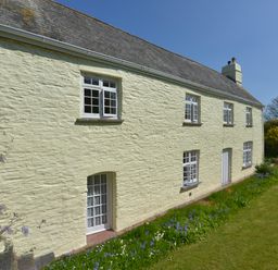 An exterior view of a two-story stone farmhouse with multiple white-framed windows and a garden with grass and flowers at Tregonhawke Farmhouse in Millbrook