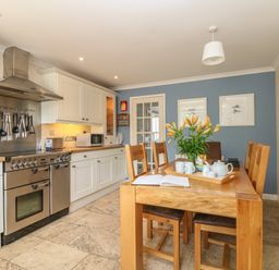 A kitchen with white cabinets a stainless steel stove and a wooden dining table with chairs and flowers at 1 River Cottage in Calstock