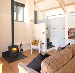 A living room with a wood burning stove a brown sofa a white chair and kitchen cabinets at Mugwell Barn Luckett near Callington