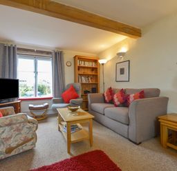 A living room with a gray sofa and armchairs around a wooden coffee table with a bookshelf and window in the background at Regatta Cottage in Feock
