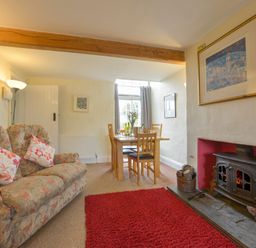 A living room with a floral sofa two pillows a red rug a wood burning stove and a dining table with chairs at Regatta Cottage in Feock