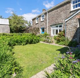 A garden with grass lawn and flowers beside a stone farmhouse with white windows and blue doors at Polcreek Farmhouse in Veryan