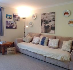 A living room with a sofa with cushions by a window with curtains and framed pictures on the wall at Stones Throw in St Mawes
