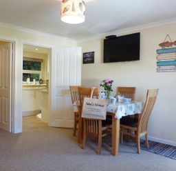 A dining area with a wooden table and chairs a wall mounted television and an open door to a kitchen at Stones Throw in St Mawes