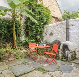 An outdoor patio with orange table and chairs surrounded by plants and a white wall with a small fireplace and a bench at Primrose Cottage Penryn