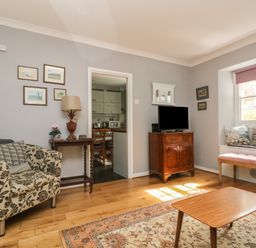 A living room with a floral armchair a wooden cabinet with a television a window seat with cushions and a wooden table at Primrose Cottage in Penryn